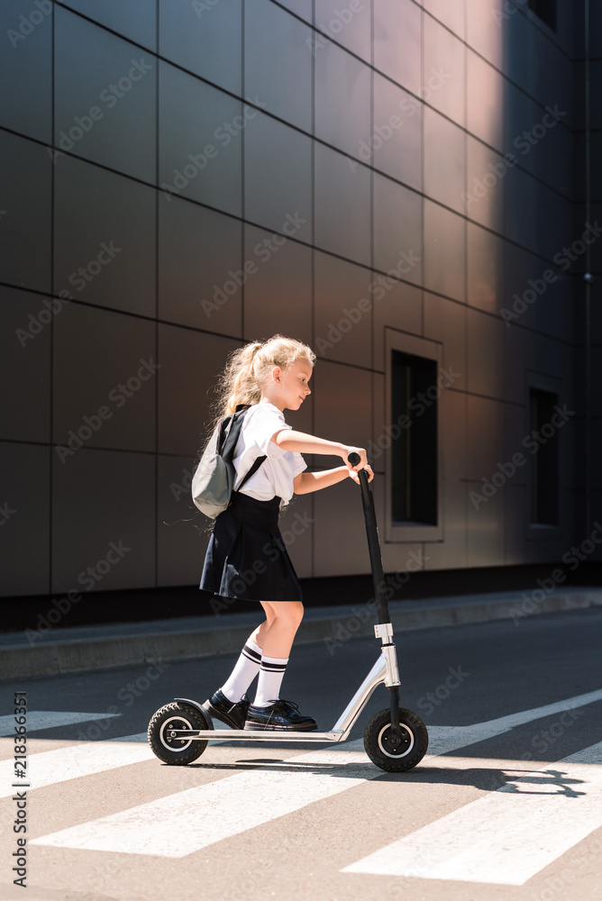 side view of cute little schoolgirl with backpack riding scooter on street side view of cute little schoolgirl with backpack riding scooter on street
