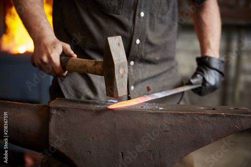 An artisan metal worker in ear protectors using a hammer to shape a red hot piece of metal on an anvil.  photo