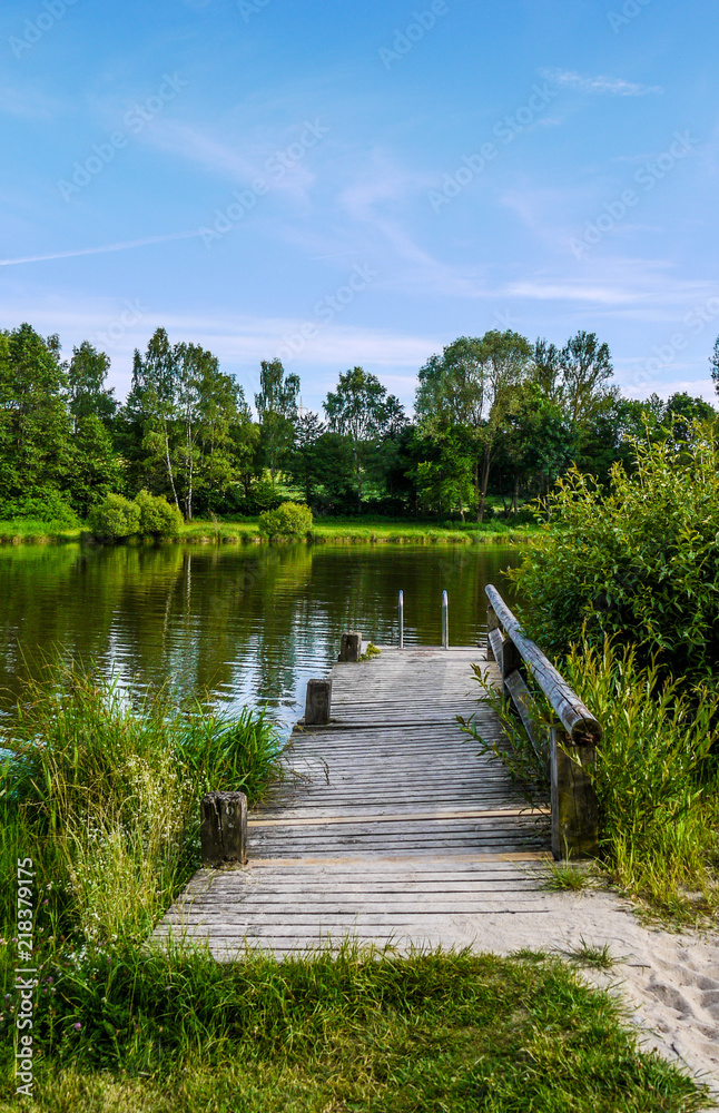 Fototapeta premium One wooden pier at the edge of a reflective lake surrounded by trees. Taken on a sunny day in Dinkelsbühl, Bavaria, Germany