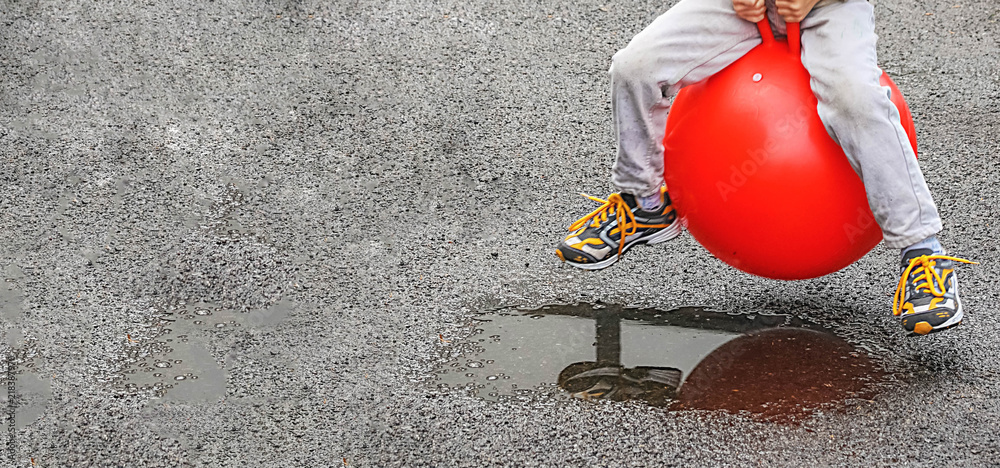 Kind bei nassem Wetter auf rotem Hüpfball im Breitbild