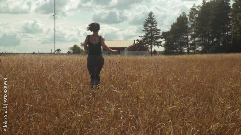 Happy brunette girl running along golden field toward country house and trees in bright sunny day. A young woman in black t-shirt and trousers runs in slow motion rear view. Happy brunette girl running along golden field toward country house and trees in bright sunny day. A young woman in black t-shirt and trousers runs in slow motion rear view.