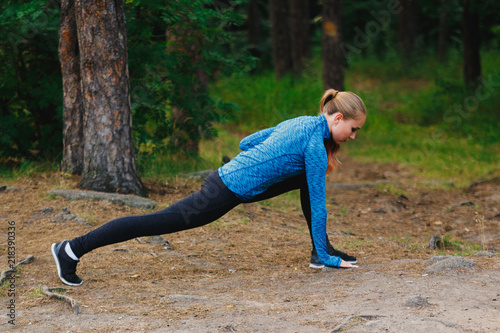 Legs stretching in low lunge. Girl in blue sportswear doing exercises outdoors in coniferous forest. Healthy lifestyle sport concept.