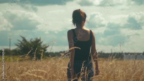 Sad girl walks along the golden field in a bright sunny day. Young brunette woman in black t-shirt and pants walking in slow motion rear view