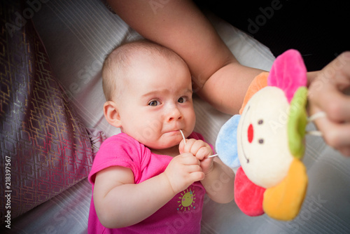 Sweet adorable baby girl puts string to mouth looking towards camera. 6-7 months old infant lying on back with colorful toy