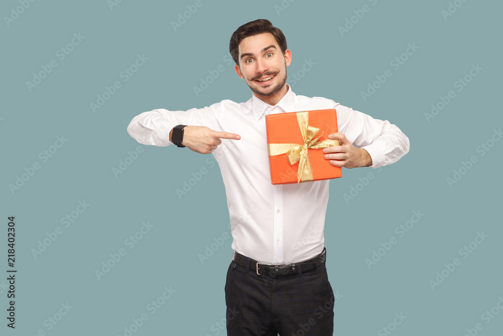 Funny happy bearded man in white shirt standing and holding red gift box, pointing finger at present and looking at camera with toothy smile. Indoor, studio shot isolated on light blue background Funny happy bearded man in white shirt standing and holding red gift box, pointing finger at present and looking at camera with toothy smile. Indoor, studio shot isolated on light blue background