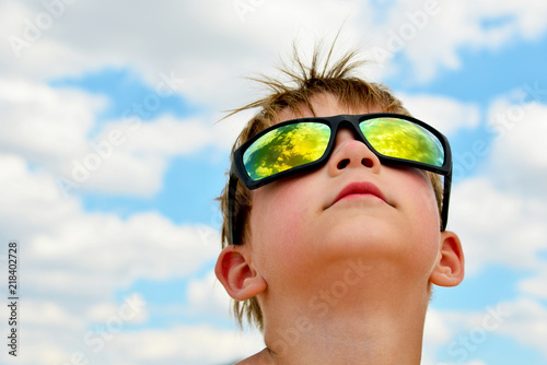 A little boy looks at the cloudy sky with glasses. The clouds are reflected in the glass. Yellow glasses against the blue sky.