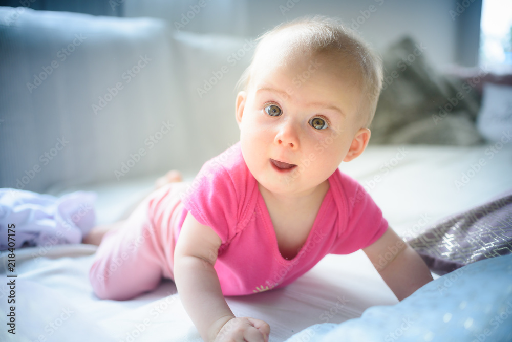 Fototapeta premium Sweet adorable baby girl lying on a couch looking towards camera. 6-7 months old infant on belly lifting upper body