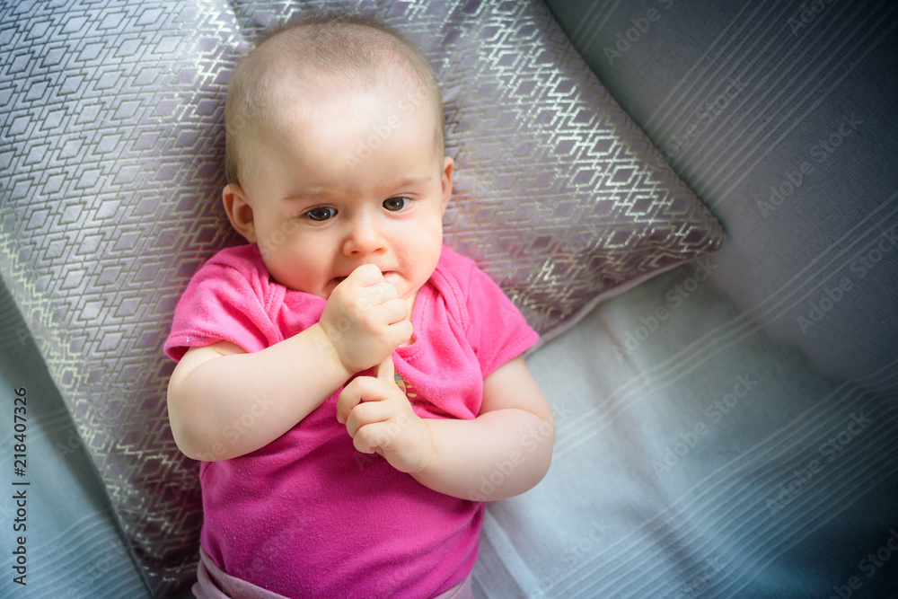 Fototapeta premium Sweet adorable baby girl puts fingers to mouth looking towards camera. 6-7 months old infant lying on back