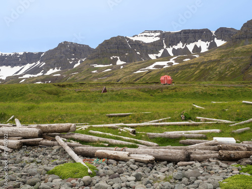 Hloduvik campsite with Red emergency shelter cabin standing on the grass meadow with view on snow coved mountain cliffs, stone and wodd logs, Hornstrandir, west fjords, Iceland photo