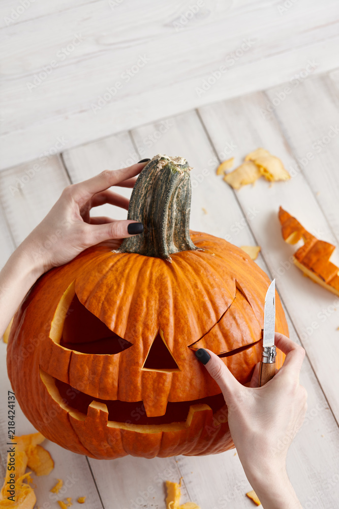 Obraz premium Woman carving big orange pumpkin into jack-o-lantern for Halloween holiday decoration on white wooden planks, close up view