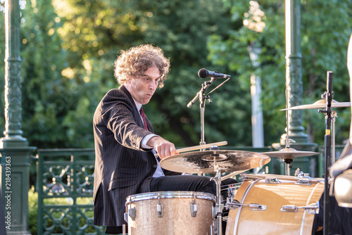 Attractive forty something man plays drums in a band, outside in a park, on a gazebo stage.