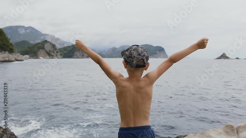 Back view. Little boy 7-8 years rejoices in life by raising his hands up standing on a rock by the sea. Vacation on the Adriatic sea. Summer trip, vacation on the sea photo