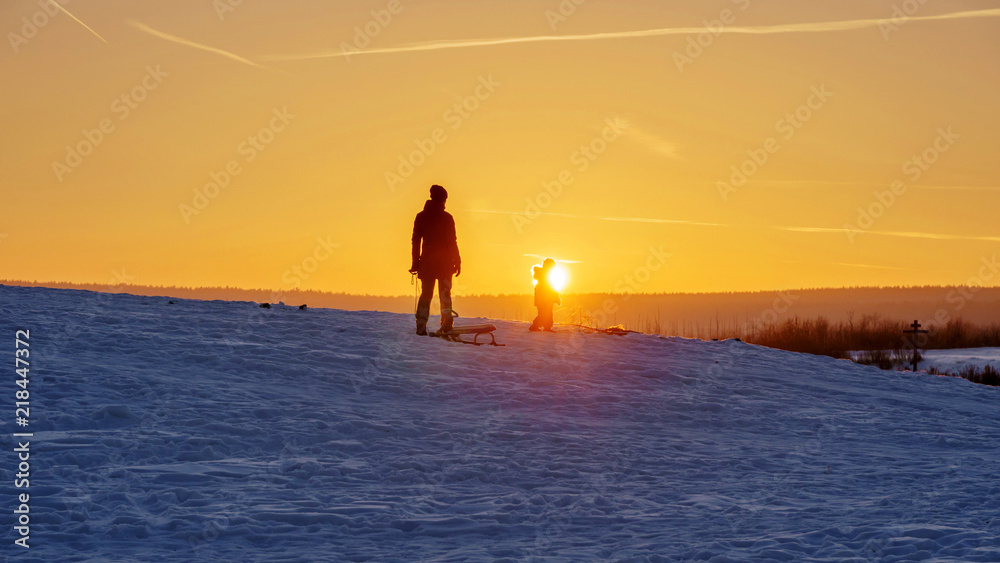 a woman with a child walks at sunset in winter with sleds