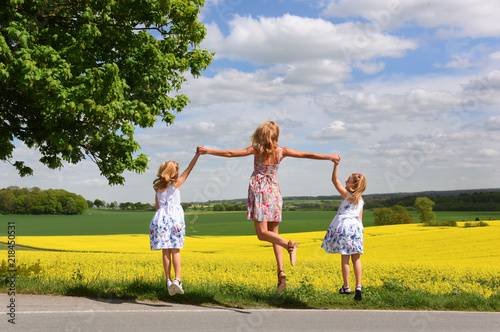 A young mother and her twin daughters jump for joy at the scenic view of the rolling landscape and yellow rapeseed fields that lay  picturesque in front of them.