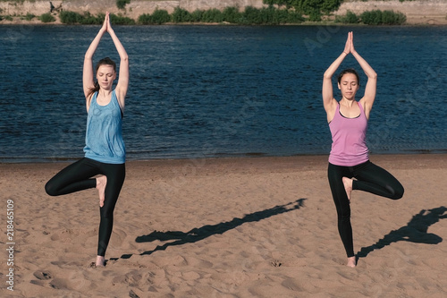Two woman stretching yoga standing on the beach by the river in the city. Beautiful city view. Balance pose. photo