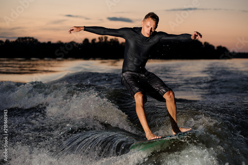 Active wakesurfer in black swimsuit jumping on a wake board at the summer evening