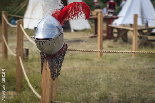 Fototapeta Naklejka Na Ścianę i Meble -  Metal knightly helmet with feathers on a blurred background of a medieval military camp. Reconstruction of battles.