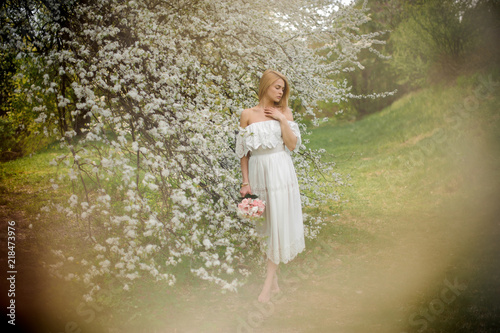 Tender full height portrait od a blonde woman in white dress with a bouquet standing near the blooming tree on misty day