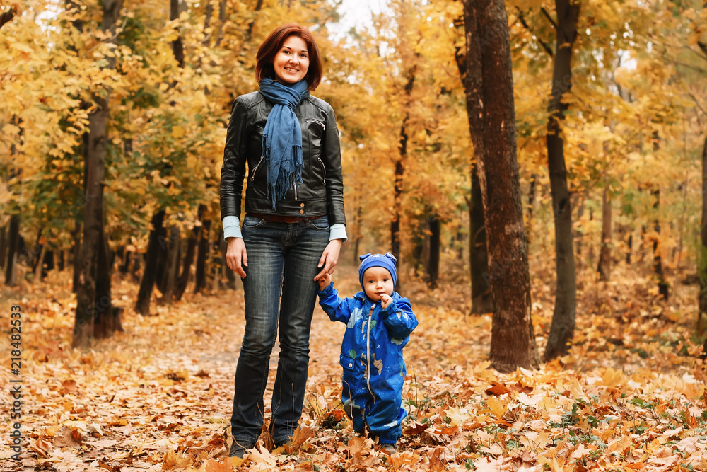 Portrait of a mother with little son in blue jumpsuit walking at autumn forest or park along the pathway covered with fallen leaves. Mom is holding hand of her toddler boy. Happy family lifestyle Portrait of a mother with little son in blue jumpsuit walking at autumn forest or park along the pathway covered with fallen leaves. Mom is holding hand of her toddler boy. Happy family lifestyle