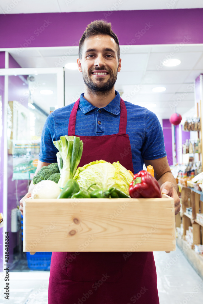 Fototapeta premium Handsome sellman holding a box with fruits and vegetables in health grocery shop
