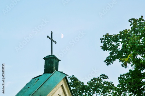 Metal christian cross on top of green roof of a church in Czech republic, Europe, blue sky with blurry white half moon in distance, green tree, rural countryside