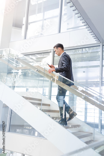 Young smiling businesss man walking up  the stairs  of a public building  and using a smartphone. Business, technology and communication concept