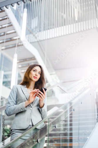 A young brunette girl in a gray jacket is standing in a bright corridor  looking into the distance and holding the phone. Business, technology and communication concept