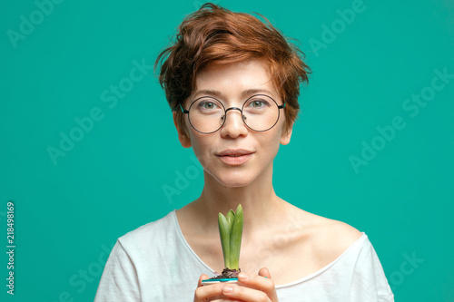 Adorable, a little bit shy hipster woman wearing spactacles and white t-shirt posing isolated with positive expression, holding in hands flower in pot photo