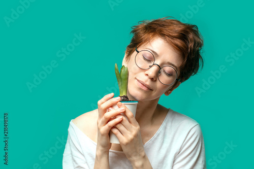 Caucasian woman with ginger hair wearing spectacles, looking and smiling at the camera posing with potted flower against blue studio background. photo