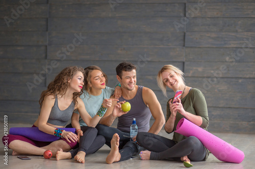 Fitness friends, man and three woman sitting on floor, having light fruit snack and watching photos on smartphone after successful yoga exercising in gym.