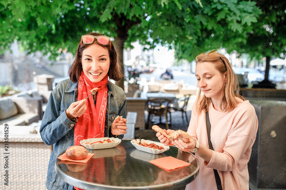 Two girl friends students eating Currywurst fast food German dish pork sausage in outdoor street food cafe