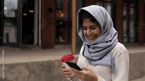 Attractive young woman in hijab walking, drinking coffee and looking at her smartphone screen. Slider real time medium shot