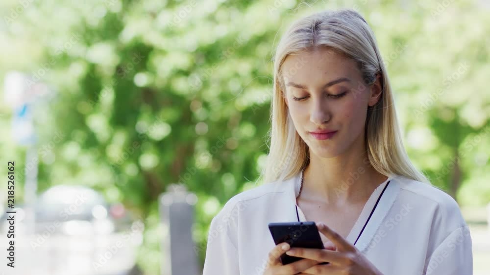 Attractive young female in white blouse browsing modern smartphone while standing on blurred background of street on sunny day Attractive young female in white blouse browsing modern smartphone while standing on blurred background of street on sunny day