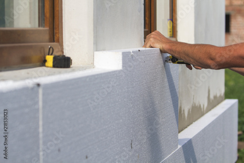 Worker hand with level and knife measuring and cutting white rigid polyurethane foam sheet on wall at newly installed plastic window. Modern technology, renovation, insulation concept.