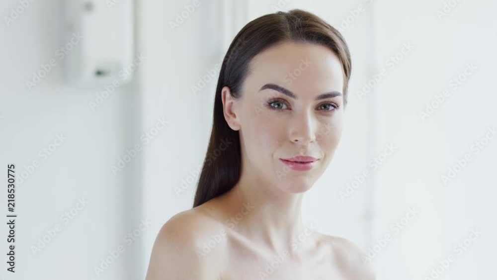 Lovely young woman with naked shoulders smiling and looking at camera while standing in white room Lovely young woman with naked shoulders smiling and looking at camera while standing in white room