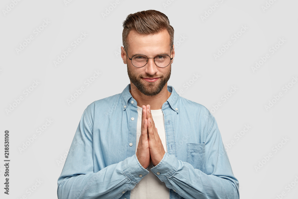 Pleased attractive unshaven man keeps hands in praying gesture, believes in good luck, has confident expression, wears round spectacles, has trendy haircut, poses indoor. People and faith concept Pleased attractive unshaven man keeps hands in praying gesture, believes in good luck, has confident expression, wears round spectacles, has trendy haircut, poses indoor. People and faith concept