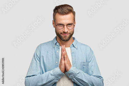 Pleased attractive unshaven man keeps hands in praying gesture, believes in good luck, has confident expression, wears round spectacles, has trendy haircut, poses indoor. People and faith concept