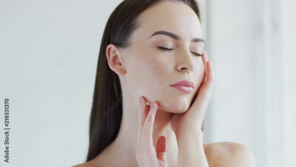 Pretty young brunette keeping eyes closed and touching face skin while standing in white room Pretty young brunette keeping eyes closed and touching face skin while standing in white room