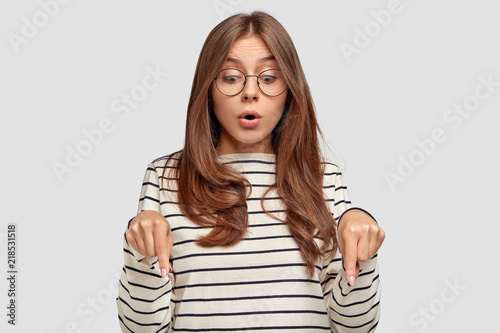 Surprised young lovely female teenager opens mouth widely, has startled look, points down with stupefied expression, sees something unbelievable, isolated over white background. Omg, look there!