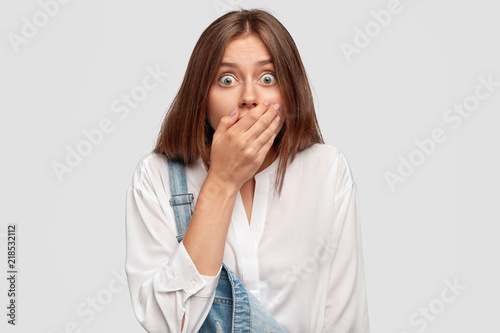 Horizontal shot of bug eyed terrific European female covers mouth, recieves awful news, dressed in fashionable white shirt and denim overalls, isolated over studio wall. People, amazement concept