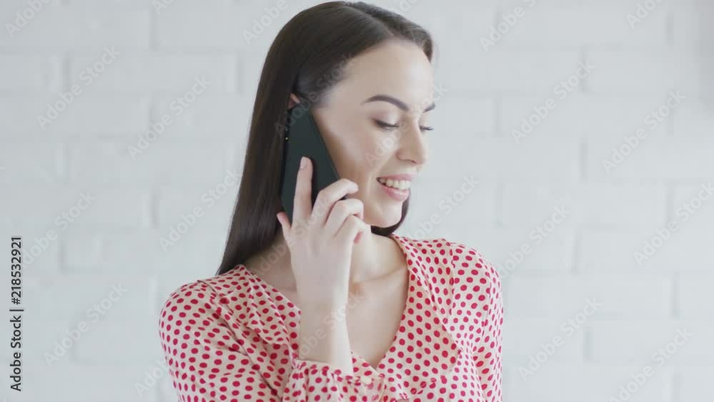 Elegant young brunette in spotted clothing smiling and talking on modern smartphone while standing near white brick wall Elegant young brunette in spotted clothing smiling and talking on modern smartphone while standing near white brick wall