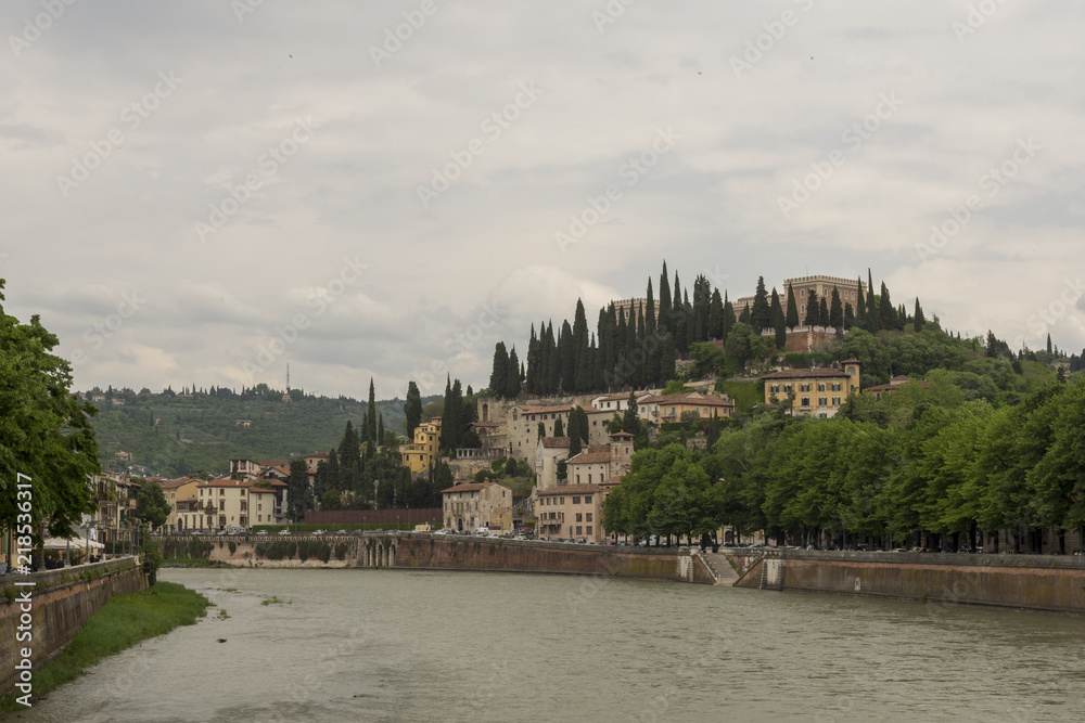 Castel San Pietro against a cloudy sky with Adige river in forefront