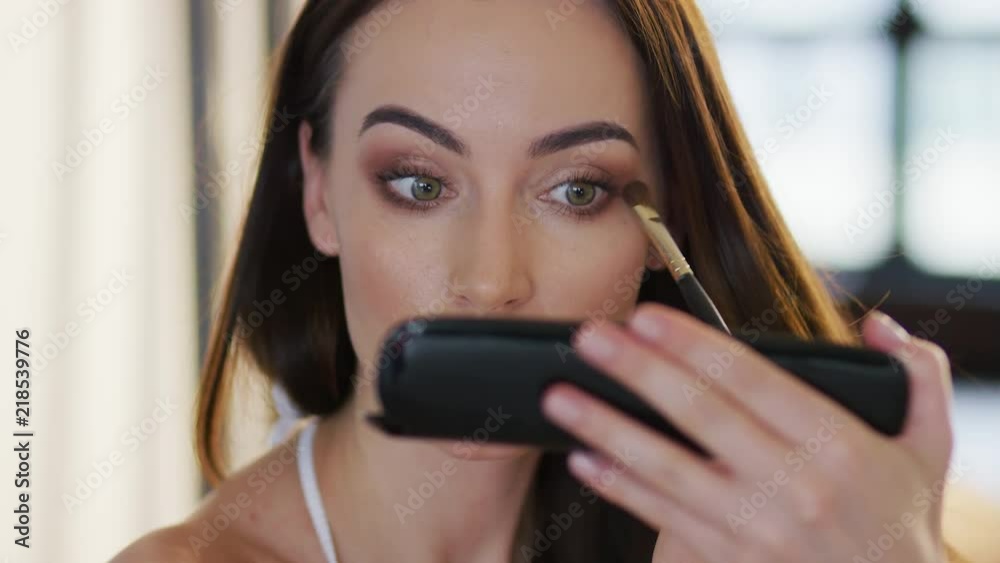Pretty young female holding makeup palette and applying eyeshadow while standing on blurred background of room