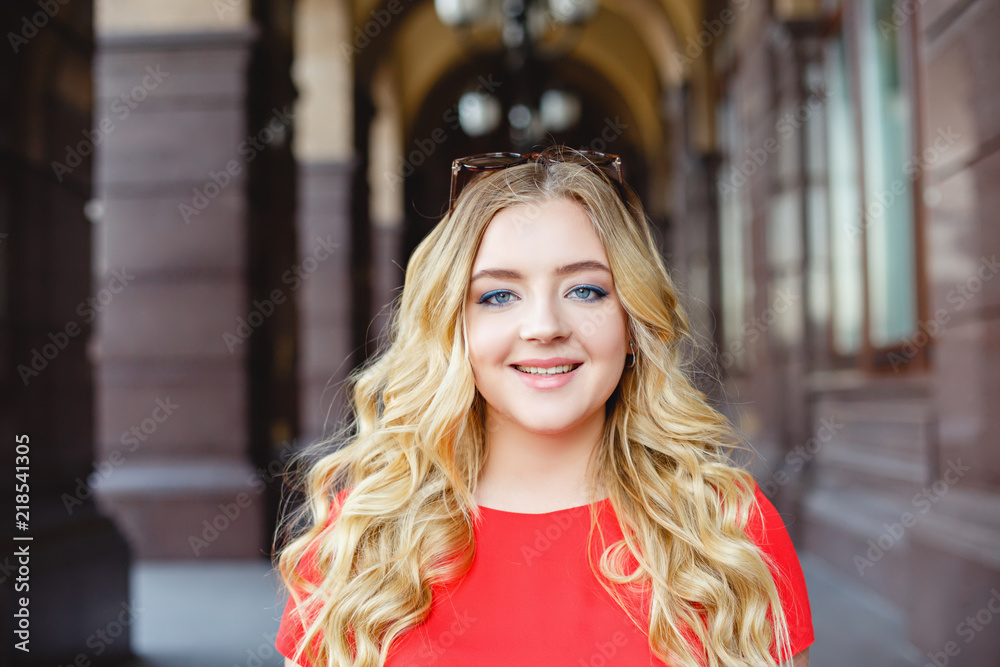Young girl on a street walk. Street style portrait. Beautiful teenager in the city. Stylish girl weared in jeans and a red T-shirt. Life style. Summer. Make up for young blonde girl. Curled blonde