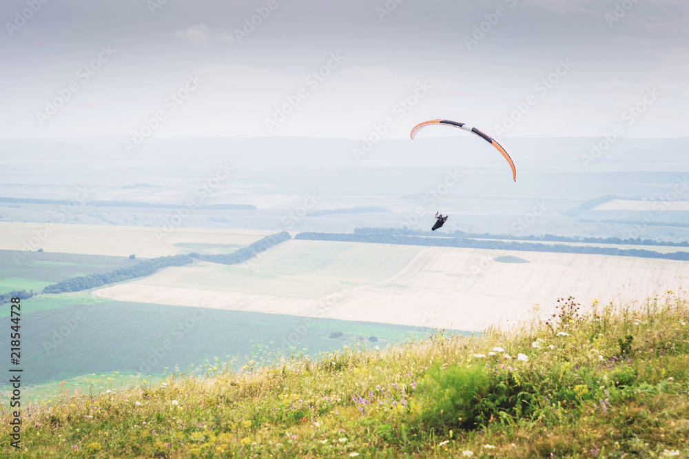 White orange paraglide with a paraglider in a cocoon against the background of fields of the sky and clouds. Paragliding Sports White orange paraglide with a paraglider in a cocoon against the background of fields of the sky and clouds. Paragliding Sports