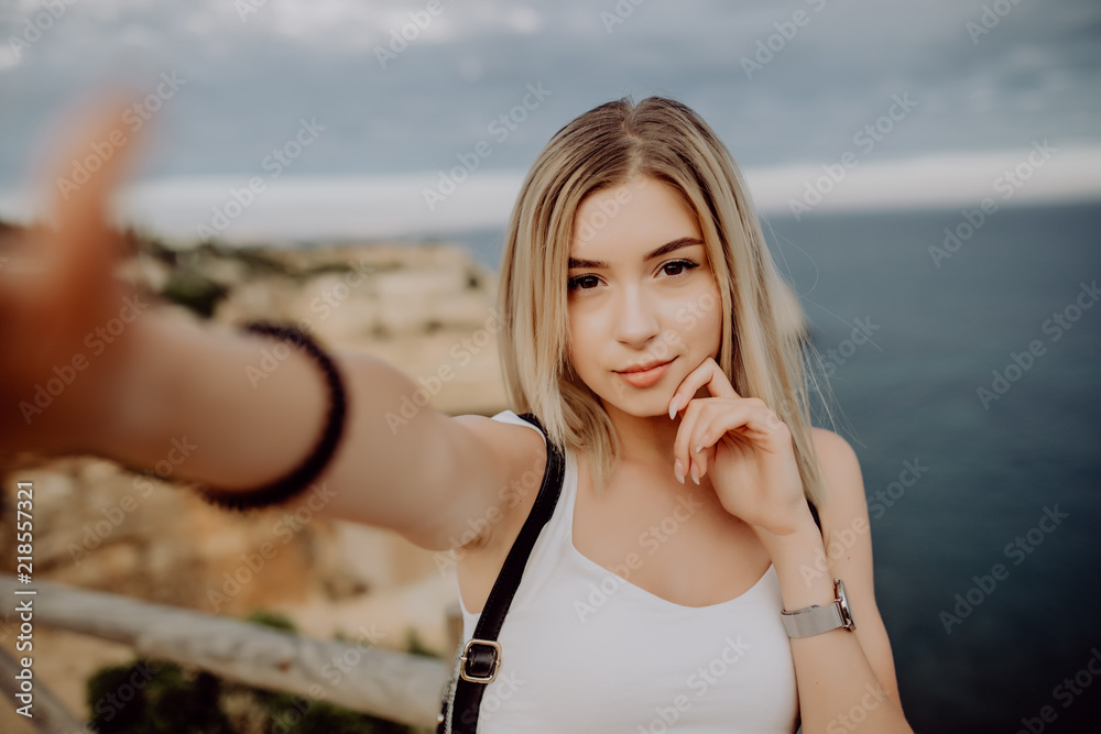 Close up shot of good looking female tourist enjoys free time outdoor near ocean on beach, looks at camera during leisure on sunny summer day, poses for selfie.
