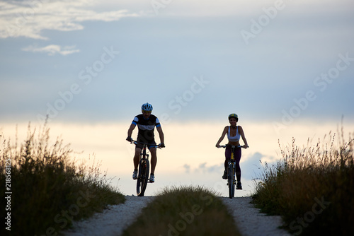 Pair of bicyclist riding bikes on a country road. Fit young people in sportswear cycling downhill against beautiful horizon with perfect sky at evening
