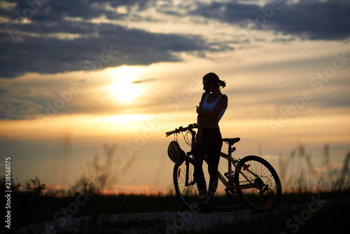 Beautiful  slender woman standing near bike and looking at sun and marvelous landscapes. Silhouette of sporty cyclist posing on trail in grass. Concept of motivation and healthy lifestyle.
