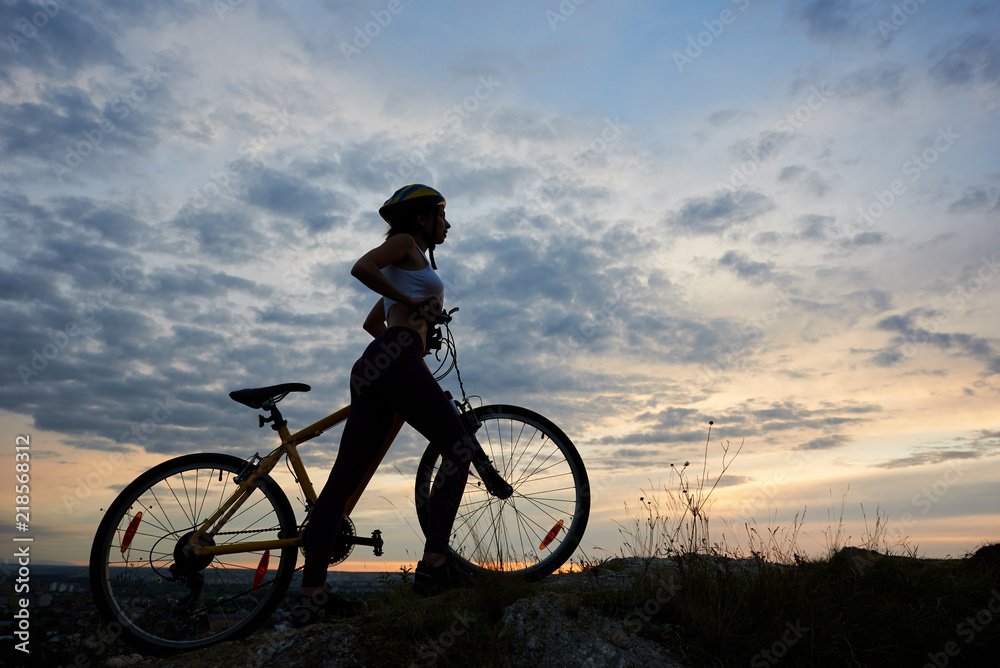Side view athletic girl in helmet with bicycle on rock under beautiful evening sky with clouds on background of city in the distance. Copy space Side view athletic girl in helmet with bicycle on rock under beautiful evening sky with clouds on background of city in the distance. Copy space