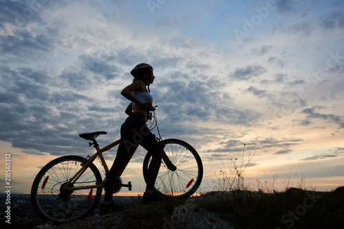 Side view athletic girl in helmet with bicycle on rock under beautiful evening sky with clouds on background of city in the distance. Copy space